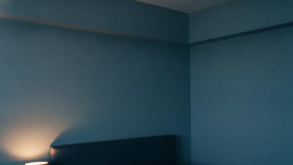 Coin de chambre le soir, cloison claire éclairée de biais par une lampe de chevet